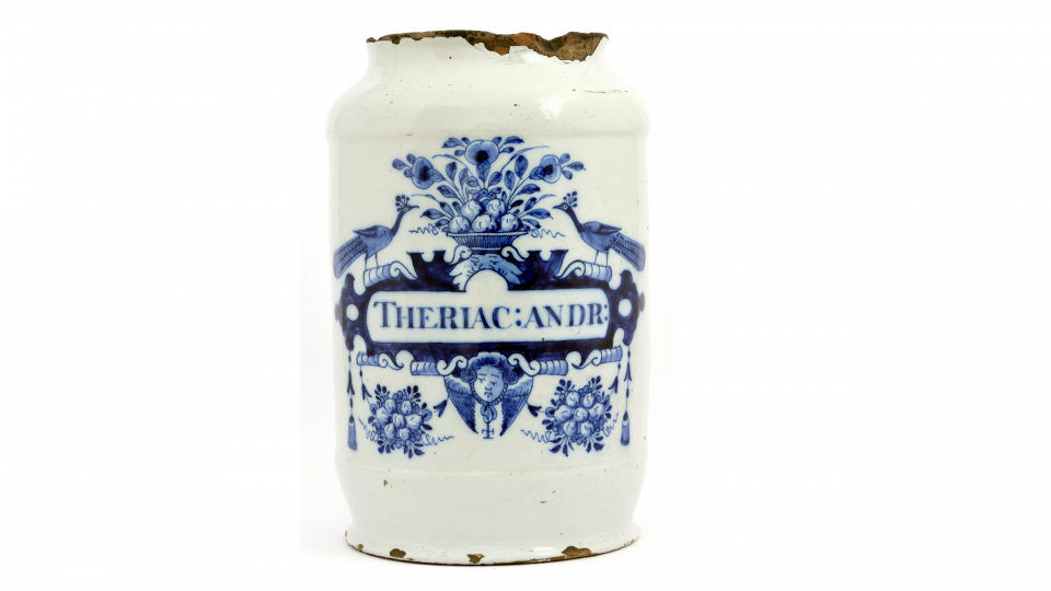 Pot de pharmacie en faïence de Delf. Blanc à motifs bleus, il présente deux ornements floraux ainsi que deux paons. L'inscription en son centre indique qu'il contient de la thériaque